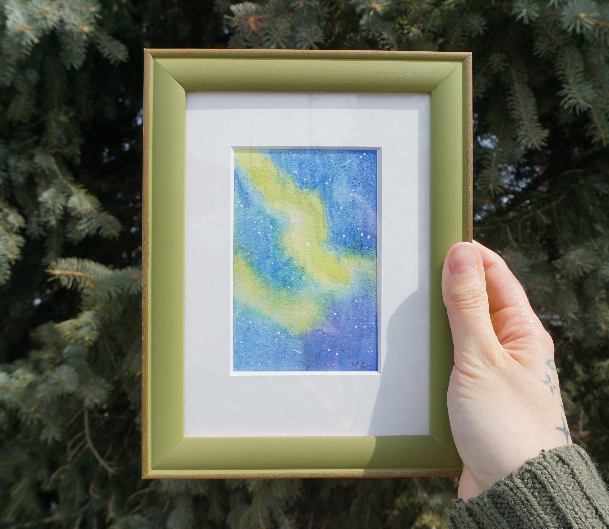 Nikki holds up their framed "Aurora Borealis 1" painting in their right hand, in front of a coniferous tree on a sunny day.