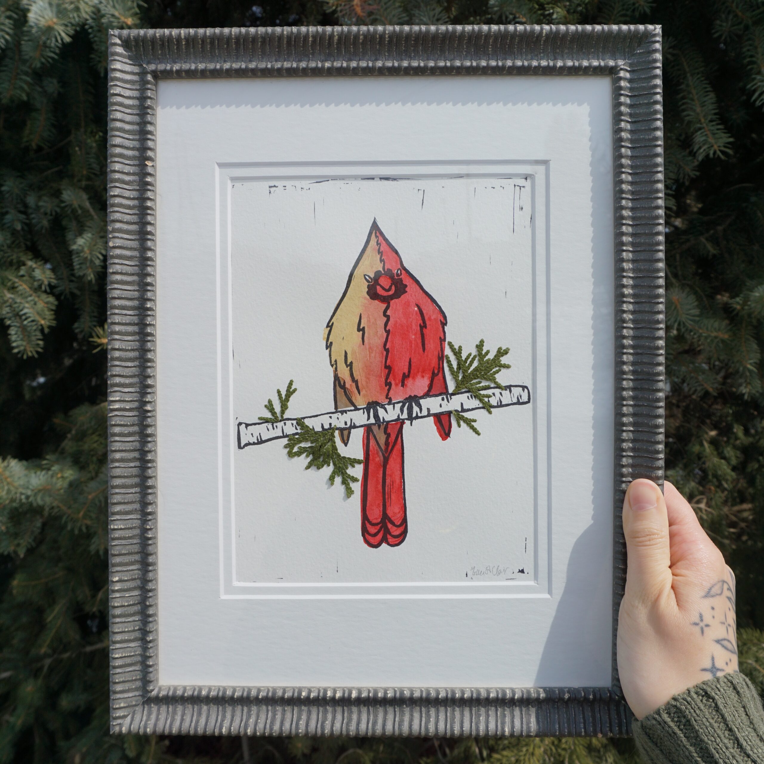 Nikki holds up their framed "Halfsider" painting in their right hand, in front of a coniferous tree on a sunny day.