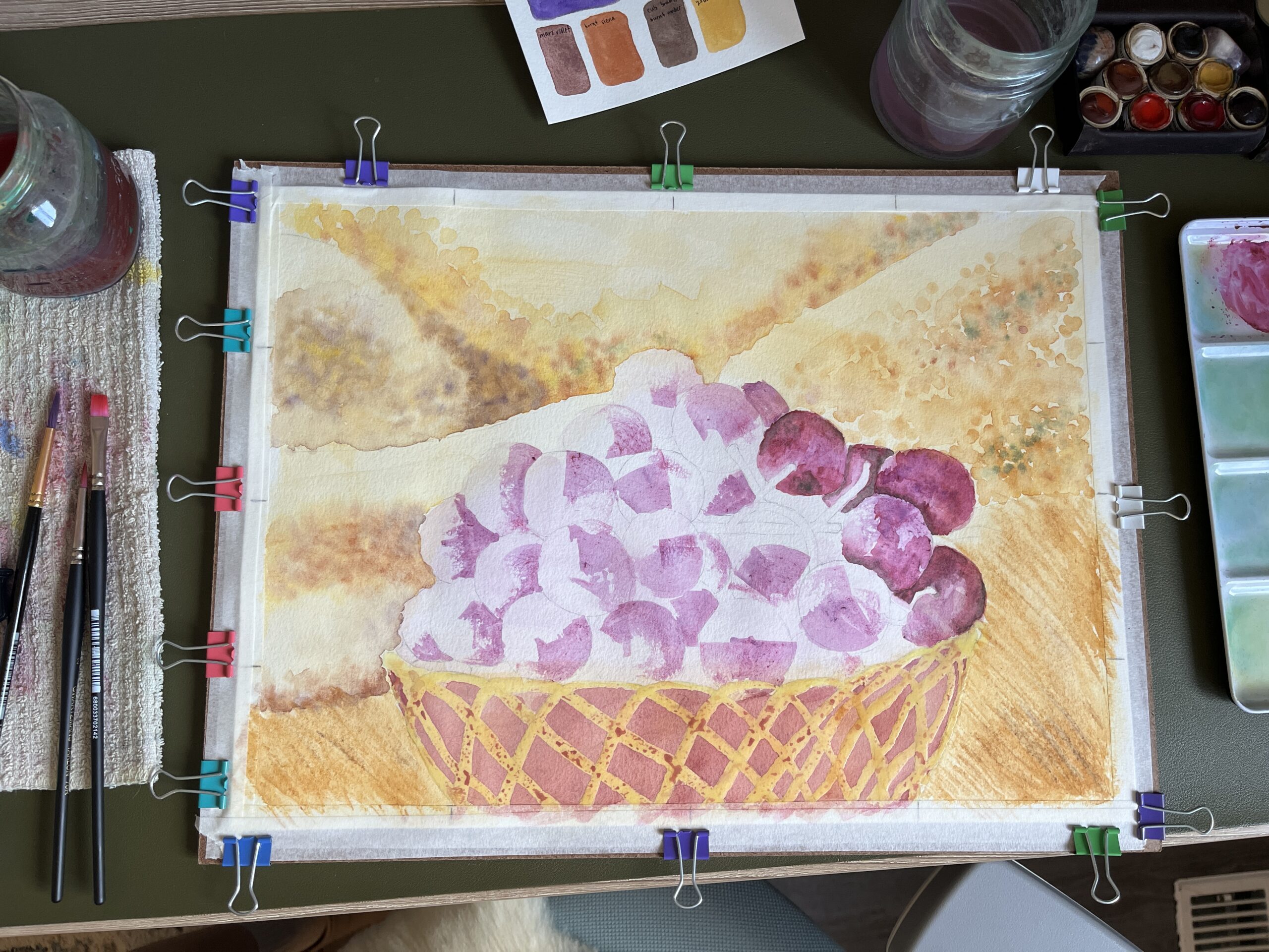 A photo of Nikki's workstation, showing their Still Life 1 painting in progress. Around the painting there is a cotton towel, water jar, paintbrushes, scrap paper, watercolour paints, and a paint mixing palette.