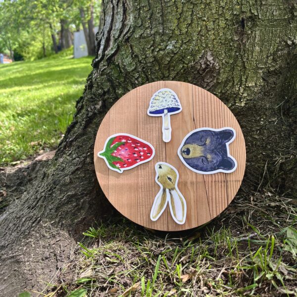 Nikki's 4 "Wheel of the Year" stickers (Mushroom, Bear, Hare, and Strawberry) displayed on a round piece of wood, which is placed on the ground at the base of a tree.