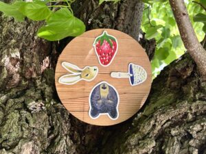 Nikki's 4 "Wheel of the Year" stickers (Strawberry, Mushroom, Bear, and Hare) displayed on a round piece of wood, which is balanced between three tree branches.