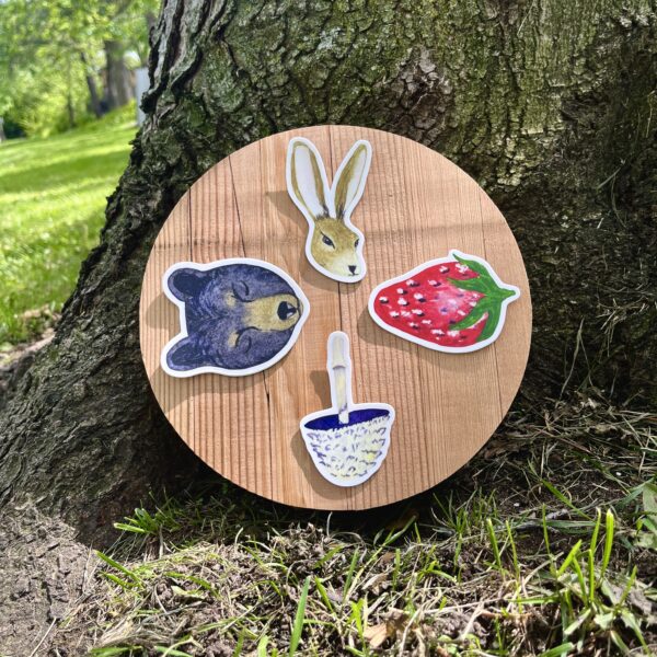 Nikki's 4 "Wheel of the Year" stickers (Hare, Strawberry, Mushroom, and Bear) displayed on a round piece of wood, which is placed on the ground at the base of a tree.
