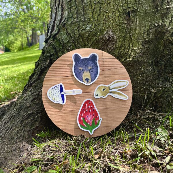 Nikki's 4 "Wheel of the Year" stickers (Bear, Hare, Strawberry, and Mushroom) displayed on a round piece of wood, which is placed on the ground at the base of a tree.