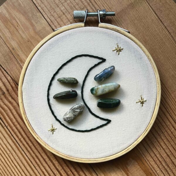 One of Nikki's "Above and Below" embroidery designs. There is a crescent moon embroidered in black thread on the left side, with 6 agate crystals (3 in the middle of the moon, and 3 to the right of it). There are three small stars embroidered around the edges of the piece in metallic gold thread.