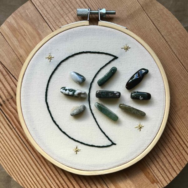 One of Nikki's "Above and Below" embroidery designs. There is a crescent moon embroidered in black thread on the left side, with 9 agate crystals (3 in the middle of the moon, and 6 to the right of it). There are four small stars embroidered around the edges of the piece in metallic gold thread.
