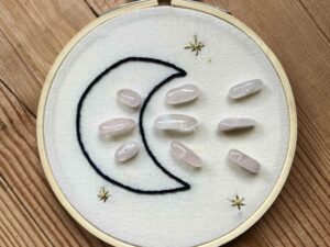 One of Nikki's "Above and Below" embroidery designs. There is a crescent moon embroidered in black thread on the left side, with 9 rose quartz crystals (3 in the middle of the moon, and 6 to the right of it). There are three small stars embroidered around the edges of the piece in metallic gold thread.