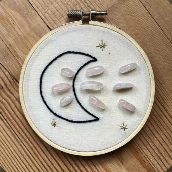 One of Nikki's "Above and Below" embroidery designs. There is a crescent moon embroidered in black thread on the left side, with 9 rose quartz crystals (3 in the middle of the moon, and 6 to the right of it). There are three small stars embroidered around the edges of the piece in metallic gold thread.