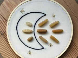 One of Nikki's "Above and Below" embroidery designs. There is a crescent moon embroidered in black thread on the left side, with 9 yellow aventurine crystals (3 in the middle of the moon, and 6 to the right of it). There are four small stars embroidered around the edges of the piece in metallic gold thread.