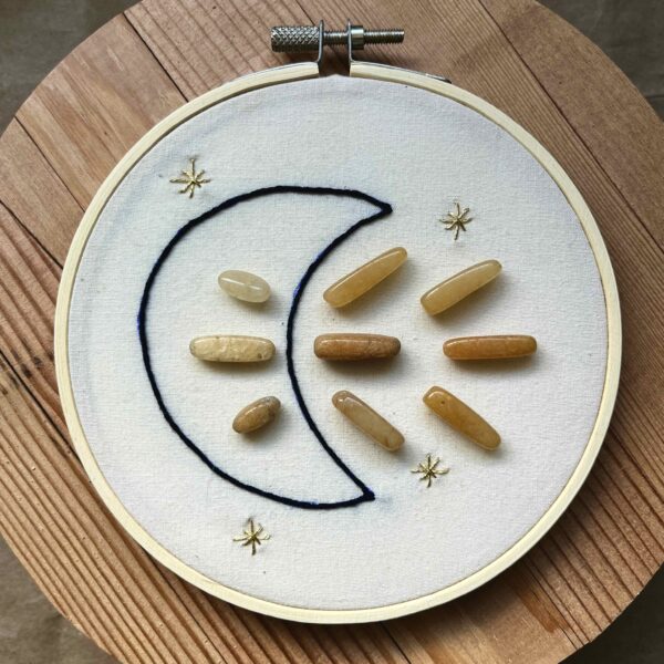 One of Nikki's "Above and Below" embroidery designs. There is a crescent moon embroidered in black thread on the left side, with 9 yellow aventurine crystals (3 in the middle of the moon, and 6 to the right of it). There are four small stars embroidered around the edges of the piece in metallic gold thread.