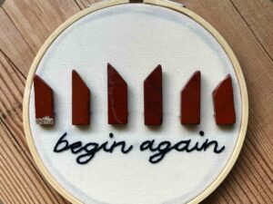 Nikki's "Begin Again" embroidery design. The words "begin again" are embroidered in black thread along the bottom, with 6 red jasper crystals arranged in a line on top of the words.