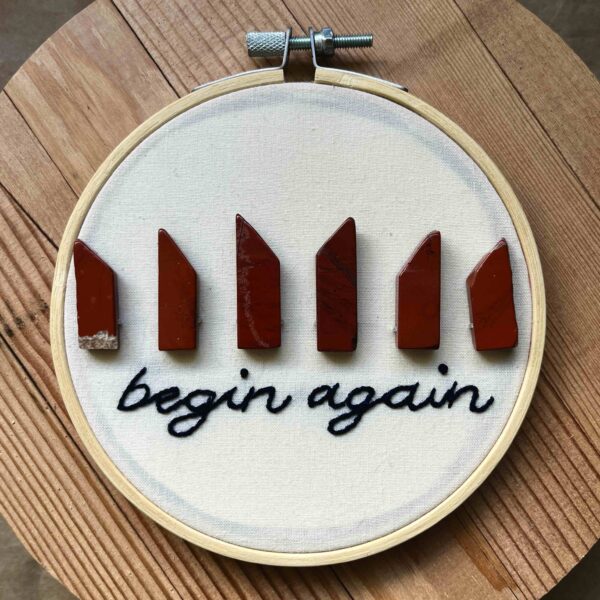 Nikki's "Begin Again" embroidery design. The words "begin again" are embroidered in black thread along the bottom, with 6 red jasper crystals arranged in a line on top of the words.