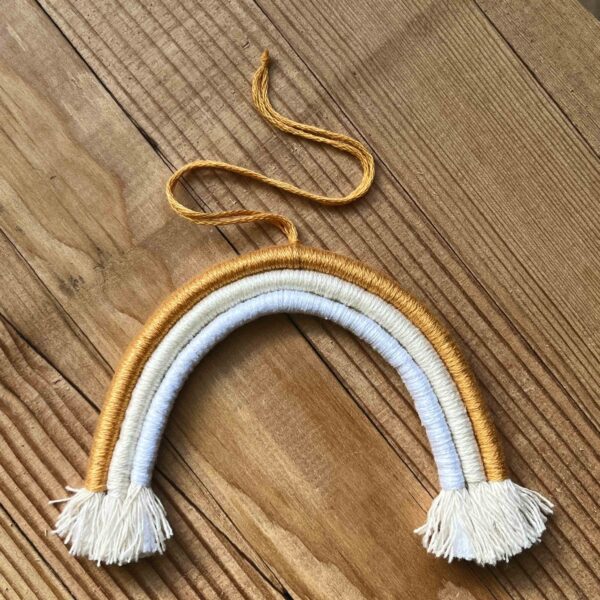 One of Nikki's mini rainbows. The rainbow is made of cotton thread, rope, and wire; and has a string to hang it from. The rainbow has three colours (listed from the outer arch to the inner one): orange, peach/light beige, white.