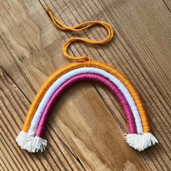 One of Nikki's mini rainbows. The rainbow is made of cotton thread, rope, and wire; and has a string to hang it from. The rainbow has three colours (listed from the outer arch to the inner one): bright orange, white, hot pink.