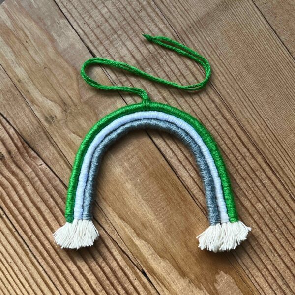 One of Nikki's mini rainbows. The rainbow is made of cotton thread, rope, and wire; and has a string to hang it from. The rainbow has three colours (listed from the outer arch to the inner one): green, white, grey.