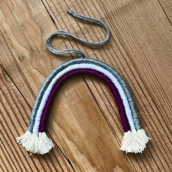 One of Nikki's mini rainbows. The rainbow is made of cotton thread, rope, and wire; and has a string to hang it from. The rainbow has three colours (listed from the outer arch to the inner one): grey, white, purple.