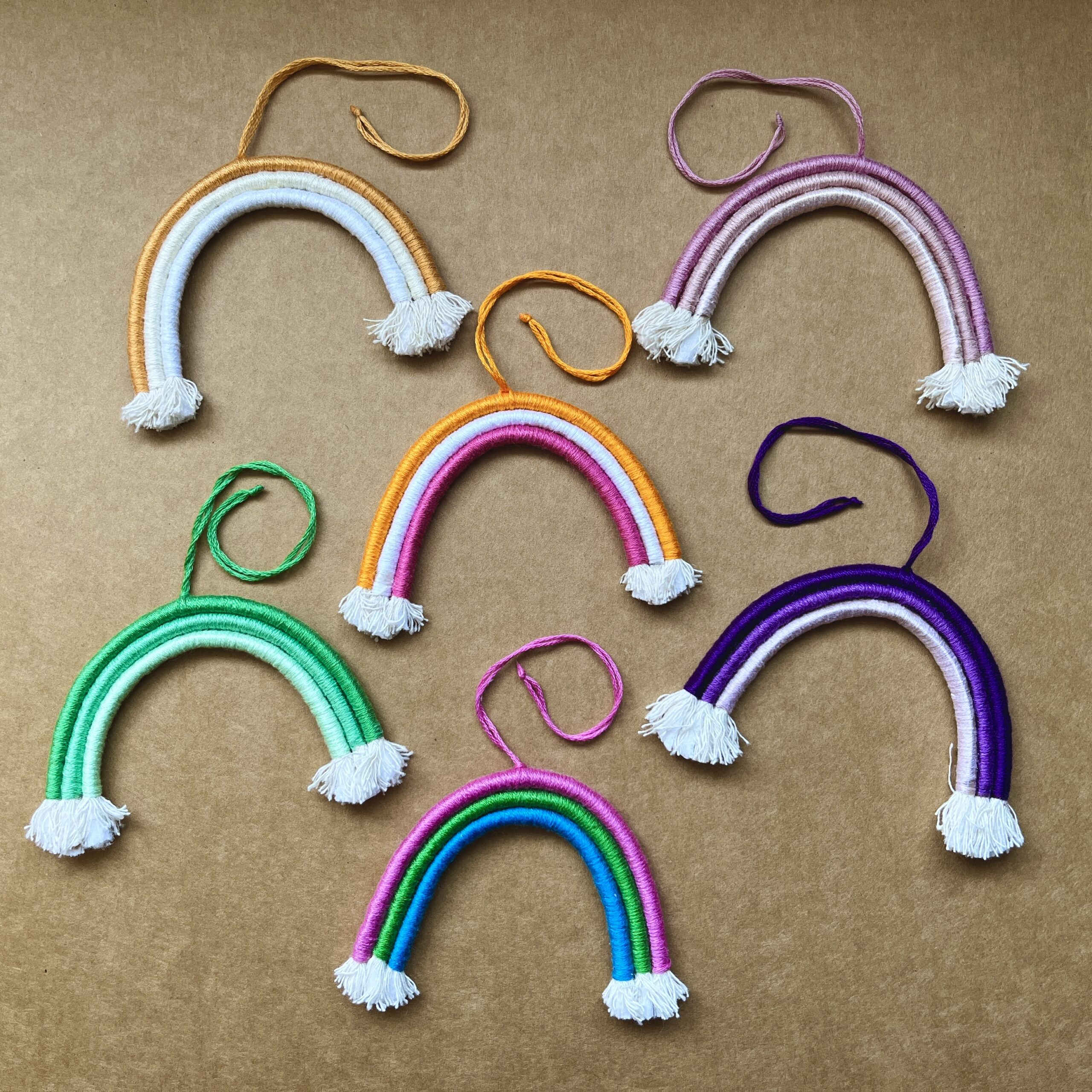 6 mini rainbows arranged on a light brown background. The rainbows are made of cotton thread, rope, and wire; and all feature a string to hang them from.